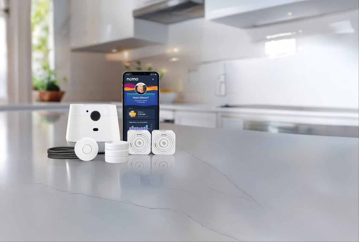 The Nomo Essential Care Kit on a kitchen counter: hub, sensors, tags, lanyard and the Nomo app showing How's Elenor?